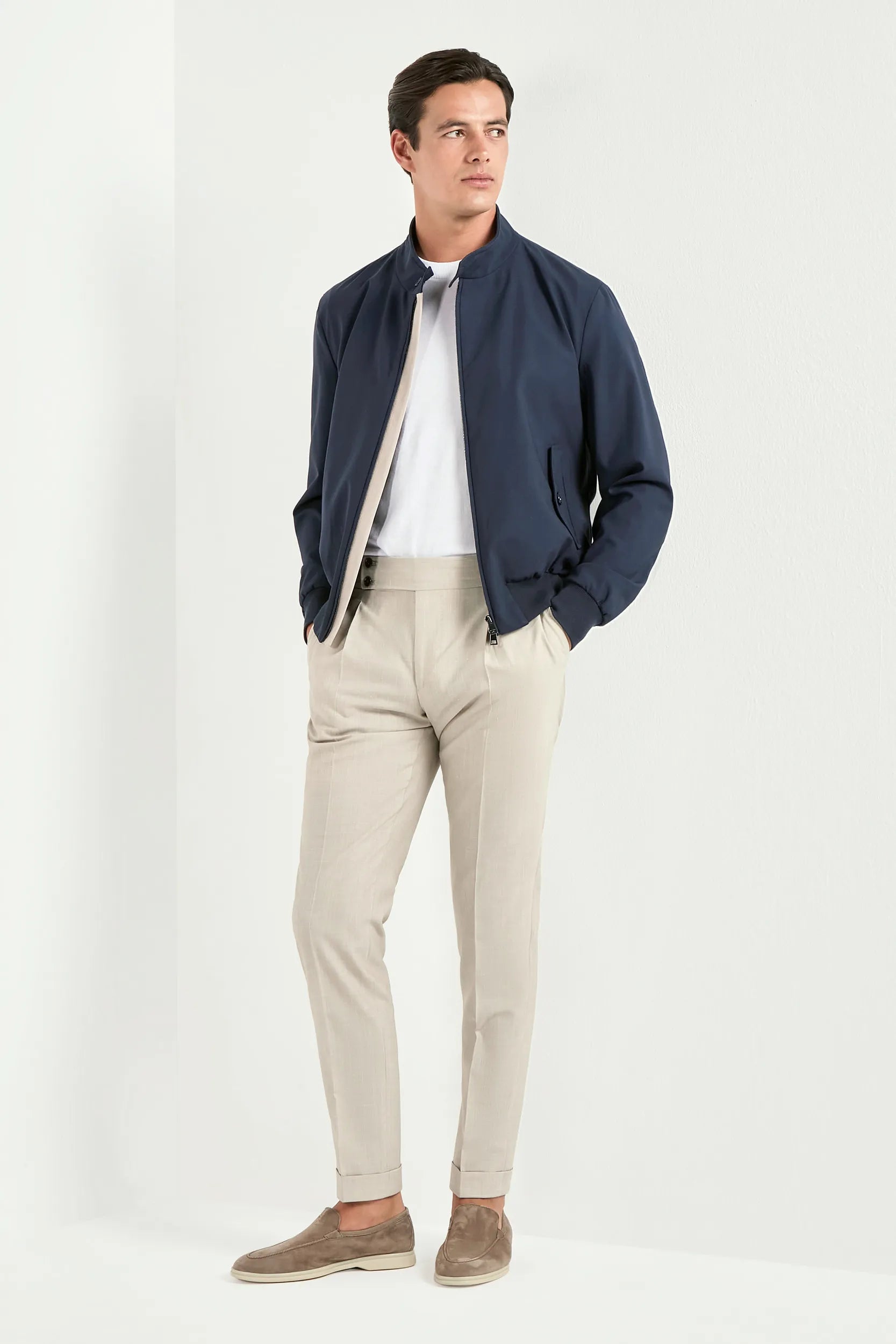 Man wearing a navy jacket, white shirt, beige pants, and brown shoes on a white background