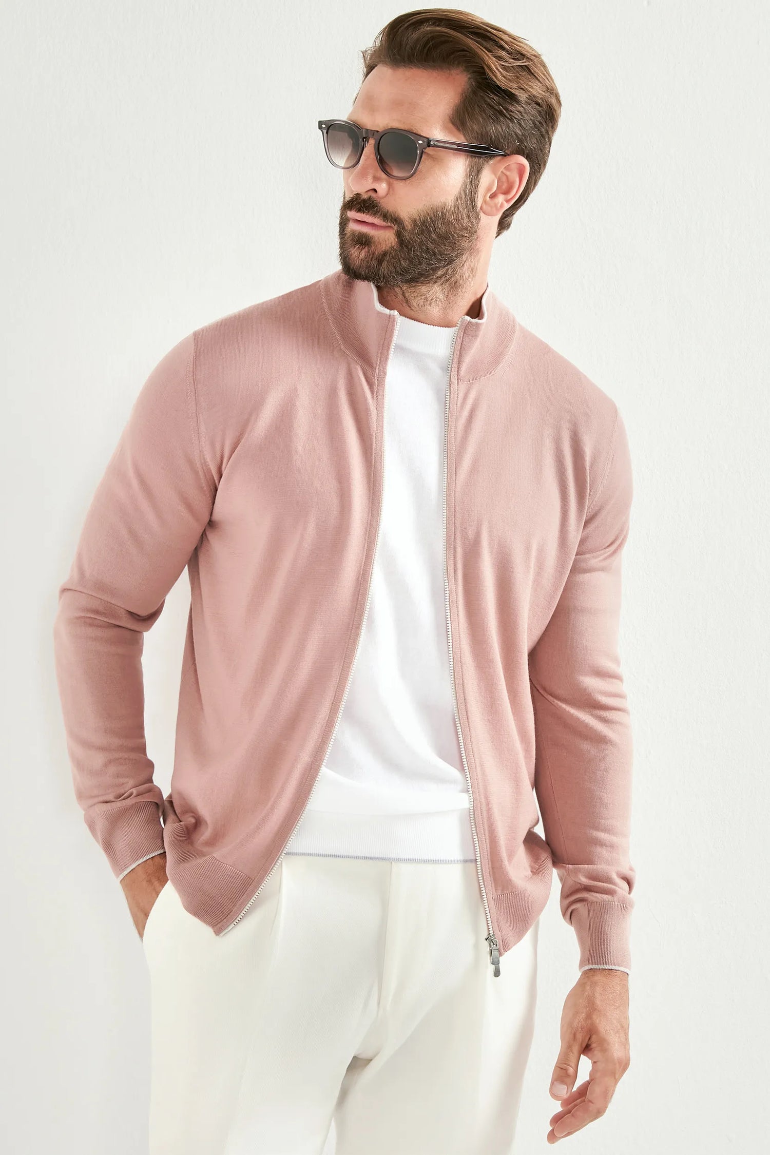 Man wearing a pink zip-up sweatshirt over a white shirt with light-colored pants on a white background