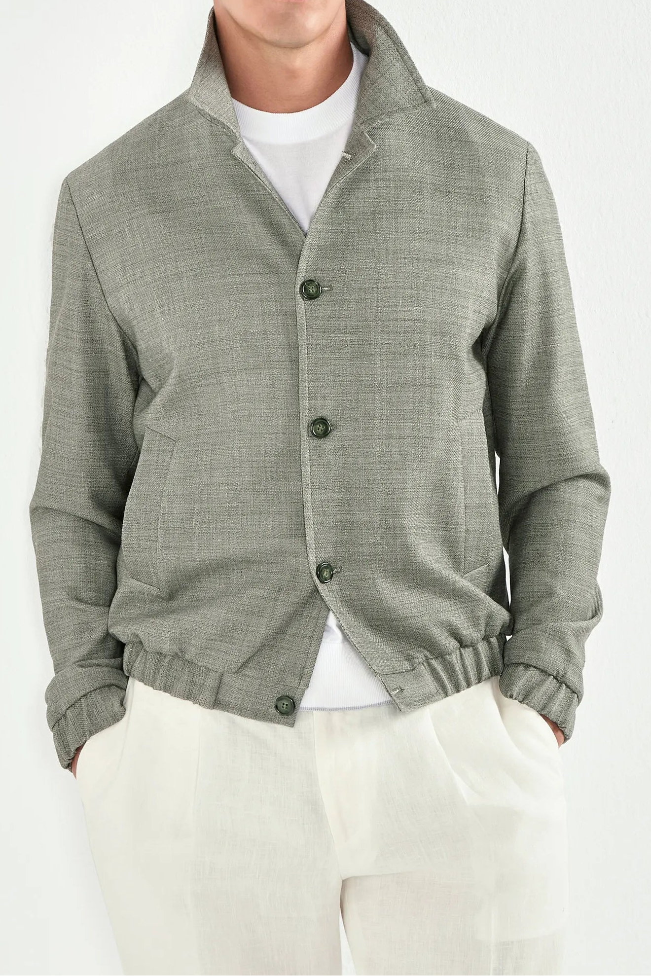 Person wearing a white t-shirt under a sage bomber jaket and white pants on a light background 