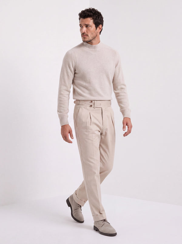 A man modeling a light beige knit mock-neck sweater and matching beige trousers with gray loafers, set against a plain white studio background.
