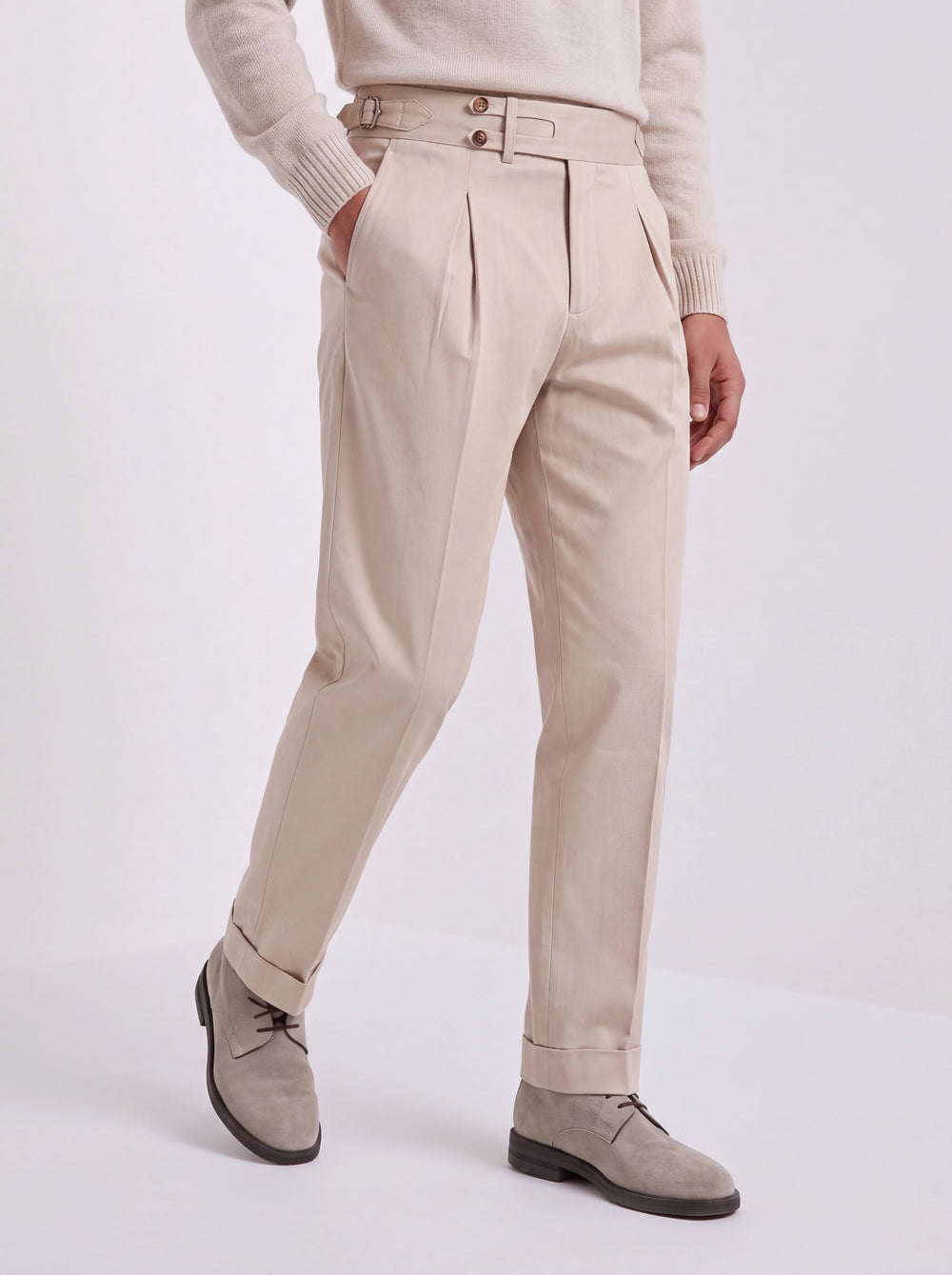 A person wearing high-waisted beige trousers with pleats and a double-breasted waist, styled with a cream sweater and taupe suede shoes against a light gray studio background.