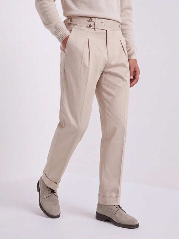 A person wearing high-waisted beige trousers with pleats and a double-breasted waist, styled with a cream sweater and taupe suede shoes against a light gray studio background.
